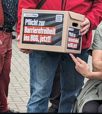Foto: zwei Hände halten eine mittelgroße Pappbox mit Deckel, auf der Box der Aufdruck von innn.it/bgg "Pflicht zur Barrierefreiheit ins BGG, jetzt! Eine dritte Hand mit lackierten Fingernägeln zeigt auf die Box