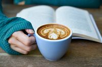 Frauenhand hält Cappucino-Tasse vor aufgeschlagenem Buch