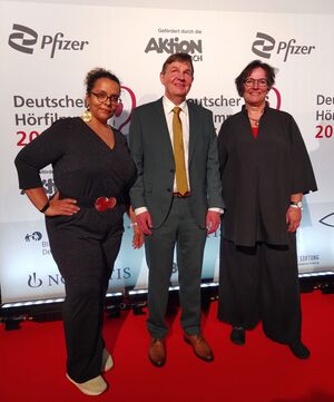 Foto: Beatrice Gómez Barroso (eine Schwarze Frau), Andreas Bethke und Martina Puschke stehen vor der Wand mit der Nennung der Sponsoren, Alle 3 tragen Abendgarderobe und sehen festlich aus