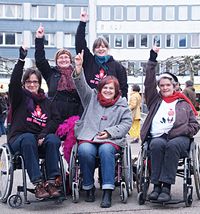 3 Weibernetz-Frauen im Rollstuhl und 2 Fußgängerinnen strecken den Arm nach oben beim One-Billion-Rising-Tanz