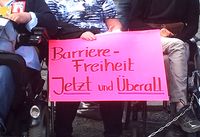 Rollstuhlfahrerinnen halten Schild mit Aufschrift: Barrierefreiheit Jetzt und Überall