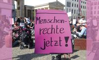 Foto einer Demo mit Menschen mit unterschiedlichen Beeinträchtigungen, stehend, im Rollstuhl, auf dem Boden sitzend; im Vordergrund ein großes handgemaltes Plakat mit der Aufschrift: Menschenrechte jetzt!