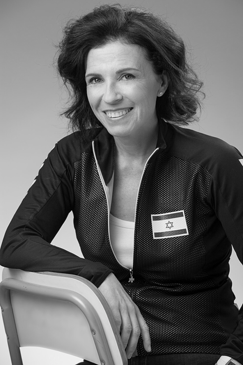 Foto: sie sitzt rittlings auf Metallstuhl, einen Arm auf Lehne. Hohe, freie Stirn, lockige, schulterlange toupierte Haare, helle Augen, Ohrstecker. Dunkle Sportjacke mit Nationalflagge von Israel und weißes T-Shirt. Gewinnendes Lächeln in die Kamera