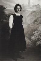 Studio-Foto: Mädchen an Schwelle zur Frau vor Landschaftsgemälde. Schwarzes langes Trägerkleid, weißer hochgeschlossener Spitzen-Pullover. Die Hände hinter dem Rücken gehalten. Dunkle Haare mit kinnlanger Frisur. Große Augen, der Blick traurig-schmollend