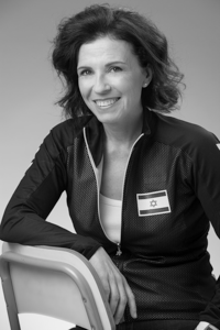 Foto: sie sitzt rittlings auf Metallstuhl, einen Arm auf Lehne. Hohe, freie Stirn, lockige, schulterlange toupierte Haare, helle Augen, Ohrstecker. Dunkle Sportjacke mit Nationalflagge von Israel und weißes T-Shirt. Gewinnendes Lächeln in die Kamera