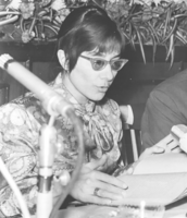 Foto:  schmales Gesicht, Frisur und Brille ganz 70er Jahre, ebenso das bunte, groß-floral gemusterte Oberteil mit großer Schleife. Sie sitzt auf Podium, in den Händen Notizbuch, liest daraus vor. Im Vordergrund Mikrofon. Im Hintergrund Zimmerpflanzen.