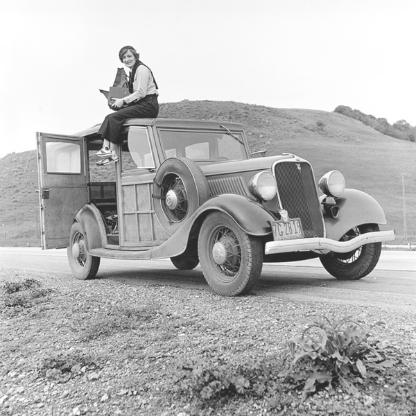 Foto: In karger Landschaft ein Oldtimer. Sie sitzt mit baumelnden Beinen auf dem Dach, hält auf dem Schoß eine große Tasche mit Ausrüstung. Hose, Bluse und Turnschuhe, den Pullover über der Schulter, auf dem Kopf eine Kappe. Blick in die Kamera.