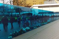 Foto der Gedenkstätte "Aktion T4 " in Berlin. Eine frei stehende große blaue Glaswand. Hinter der Wand sind Menschen, manche sitzen im Rollstuhl. Am Fuß der Wand Trauerkränze