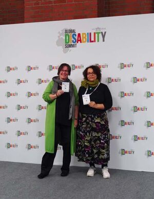 Women's network employees Martina Puschke and Beatrice Gómez stand in front of a screen with the GDS logo as a multiple pattern on it. They are smartly dressed and hold their participant badge, which is worn around their necks with a ribbon, up to the cam