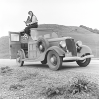 Foto: In karger Landschaft ein Oldtimer. Sie sitzt mit baumelnden Beinen auf dem Dach, hält auf dem Schoß eine große Tasche mit Ausrüstung. Hose, Bluse und Turnschuhe, den Pullover über der Schulter, auf dem Kopf eine Kappe. Blick in die Kamera.