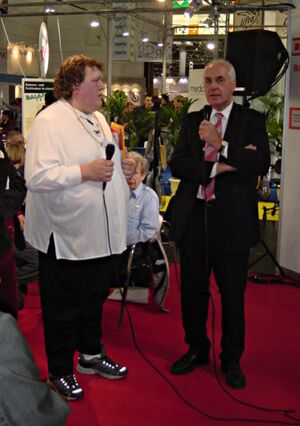 Foto: Petra Groß (lange weiße Bluse und Turnschuhe) und Karl Hermann Haack (Im Anzug) auf der Reha-Messe. Sie stehen sich gegenüber. Beide haben Mikrofone in der Hand. Im Hintergrund Messehalle mit Menschen