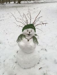 Foto: Eine Schneefrau aus drei Schneekugeln, mit zwei Brüsten. Sie trägt eine Stola aus Lebensbaumzweigen, eine Halskette aus runden Holzstücken und hat eine grüne Strickmütze auf. Die Augenbrauen aus kleinen Ästchen sind hochgezogen, der Mund wirkt freun