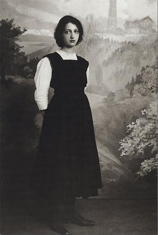 Studio-Foto: Mädchen an Schwelle zur Frau vor Landschaftsgemälde. Schwarzes langes Trägerkleid, weißer hochgeschlossener Spitzen-Pullover. Die Hände hinter dem Rücken gehalten. Dunkle Haare mit kinnlanger Frisur. Große Augen, der Blick traurig-schmollend