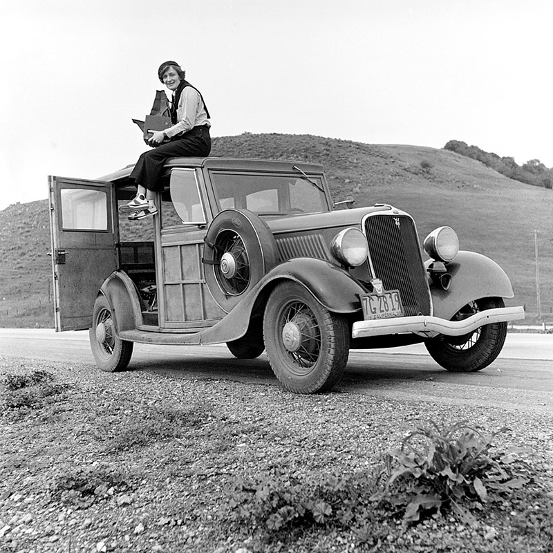 Foto: In karger Landschaft ein Oldtimer. Sie sitzt mit baumelnden Beinen auf dem Dach, hält auf dem Schoß eine große Tasche mit Ausrüstung. Hose, Bluse und Turnschuhe, den Pullover über der Schulter, auf dem Kopf eine Kappe. Blick in die Kamera.