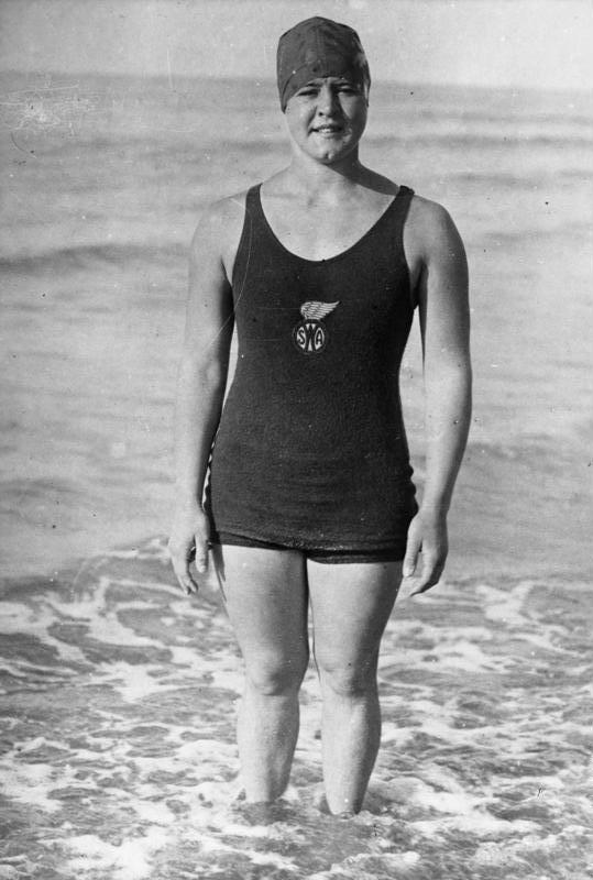 Foto: junge Frau mit eher rundem Gesicht, ausgeprägtes Kinngrübchen. Sie steht im Wasser am Meeresstrand. Enges Badedress mit langem Trägeroberteil mit Vereinslogo und Shorts sowie Badekappe. Leicht kritischer Blickt in die Kamera
