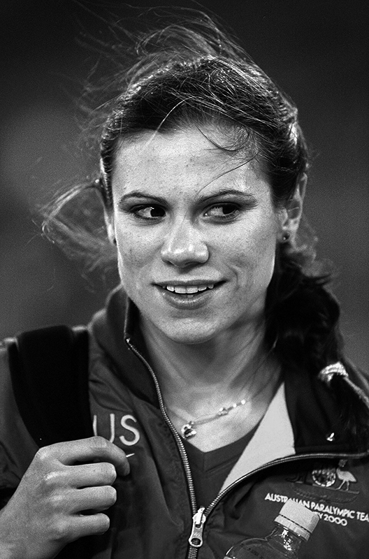 Foto: Schnappschuss- junge Frau, aus der Stirn gekämmtes, zu Zopf geflochtenes, etwas zerzaust Haar. Offizielle Paralympics-Sportjacke, eine Hand hält breiten Träger einer Tasche auf der Schulter. Blick aus dem Augenwinkel nach rechts, leicht lächelnd.