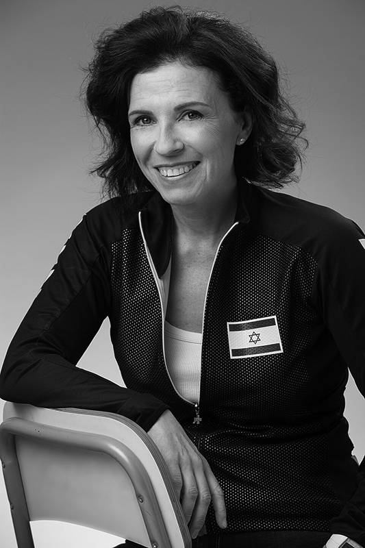 Foto: sie sitzt rittlings auf Metallstuhl, einen Arm auf Lehne. Hohe, freie Stirn, lockige, schulterlange toupierte Haare, helle Augen, Ohrstecker. Dunkle Sportjacke mit Nationalflagge von Israel und weißes T-Shirt. Gewinnendes Lächeln in die Kamera